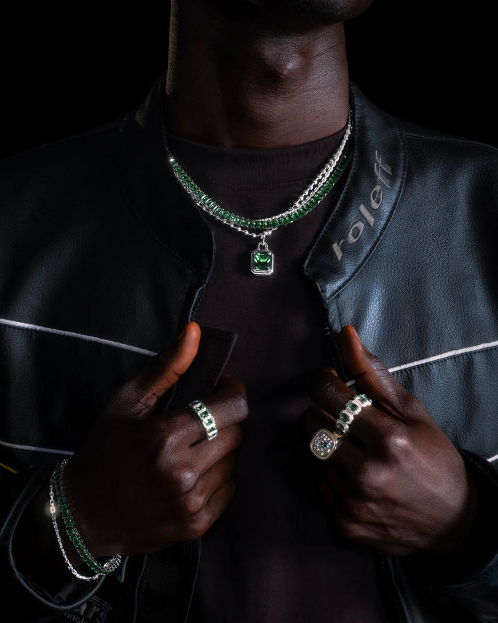 Black man wearing a black T-shirt and leather cafe racer jacket with Hatton Labs luxury silver and green necklaces, a variety of rings and precious stone bracelets.
