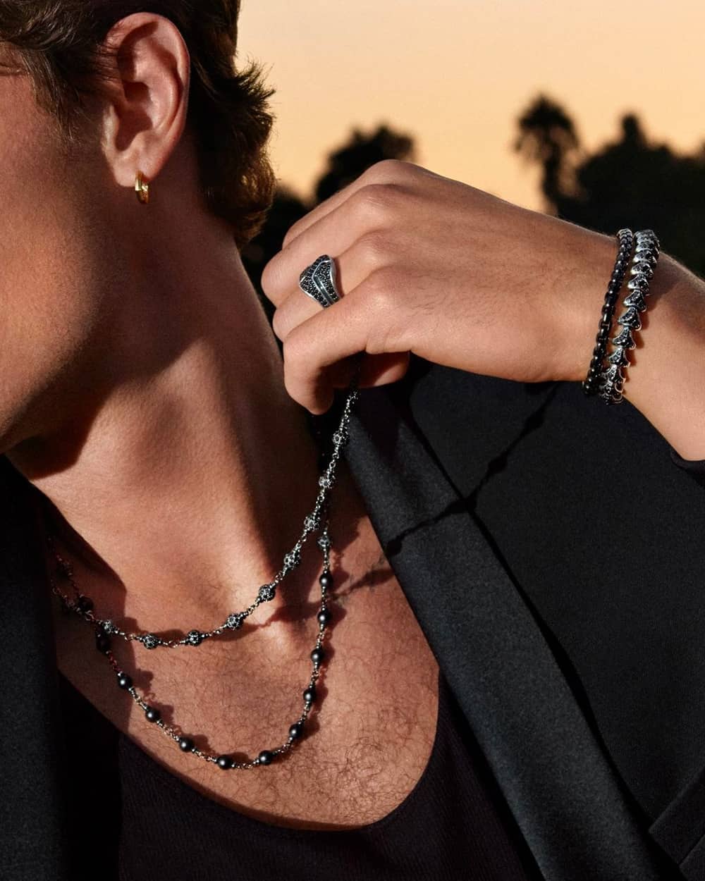 Close up of man wearing a black T-shirt and blazer, with a selection of edgy David Yurman black beaded necklaces and bracelets