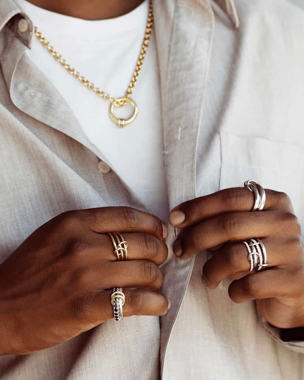 Close up of black man wearing a white T-shirt and grey shirt, with a Spinelli Kilcollin gold ring pendant necklace and multiple silver and gold rings