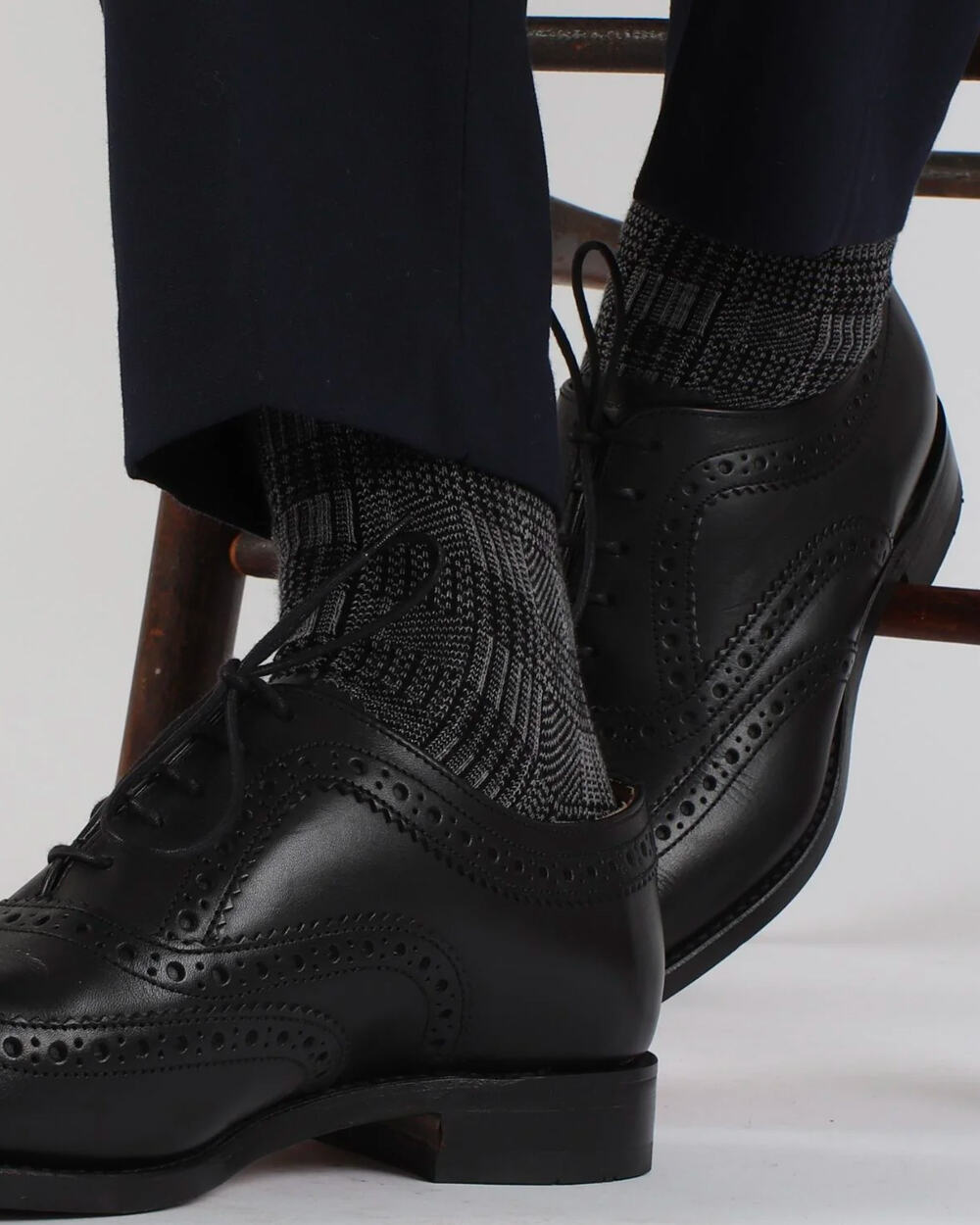 Man wearing luxury charcoal check Corgi socks on feet with navy tailored pants and black leather brogues