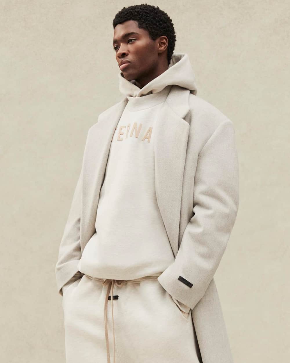 Black man wearing an off-white Fear of God hoodie, logo sweatshirt and matching sweatpants with an off-white overcoat