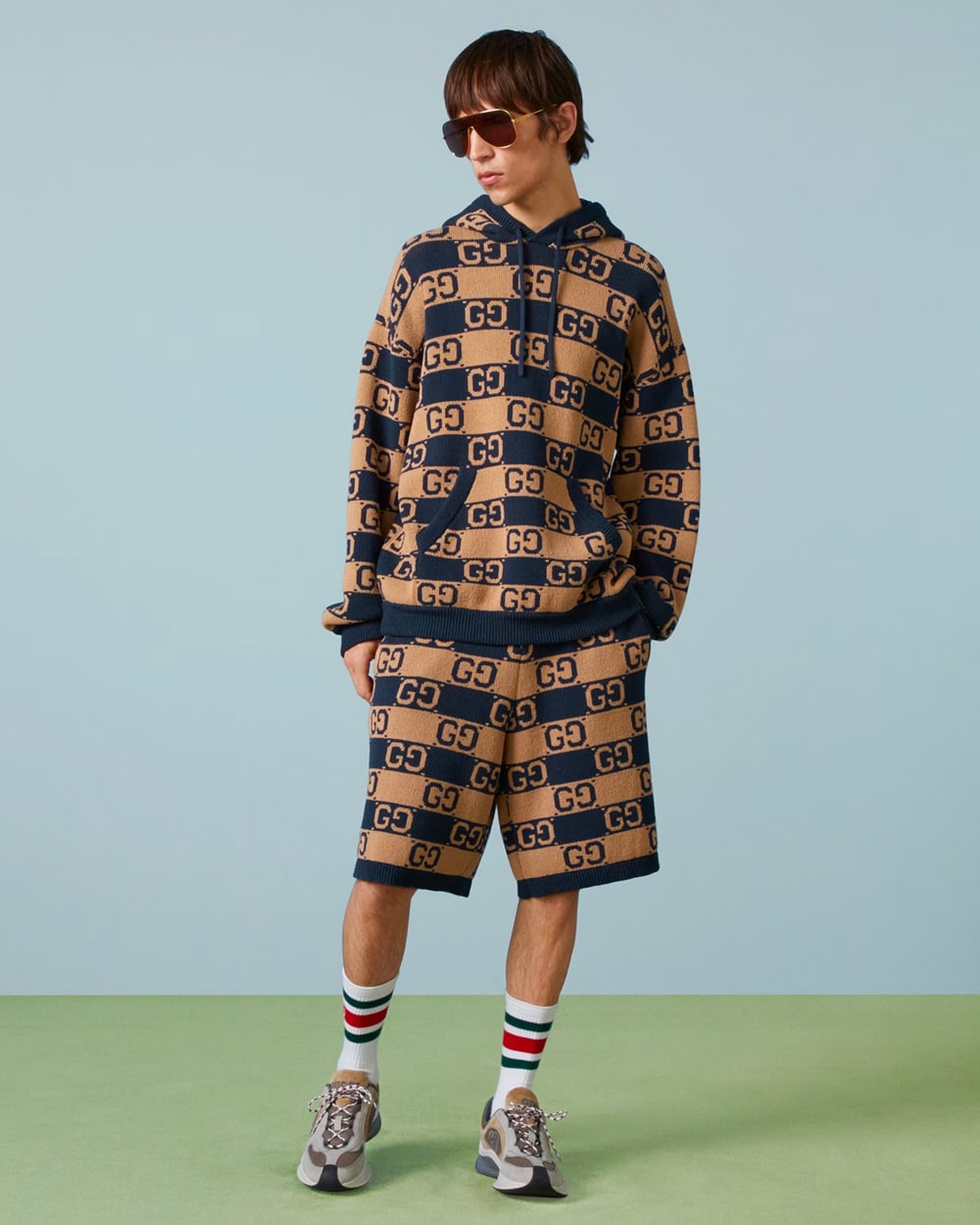 Man wearing a navy/brown check Gucci logo hoodie and matching shorts with white tube socks and sneakers