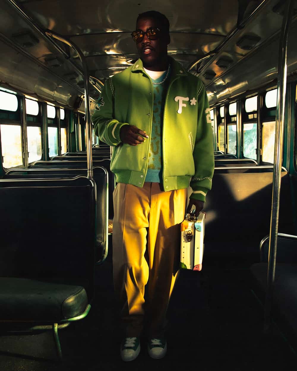 Black man wearing preppy Golf Le Fleur yellow pants, light blue v-neck sweater and green varsity jacket