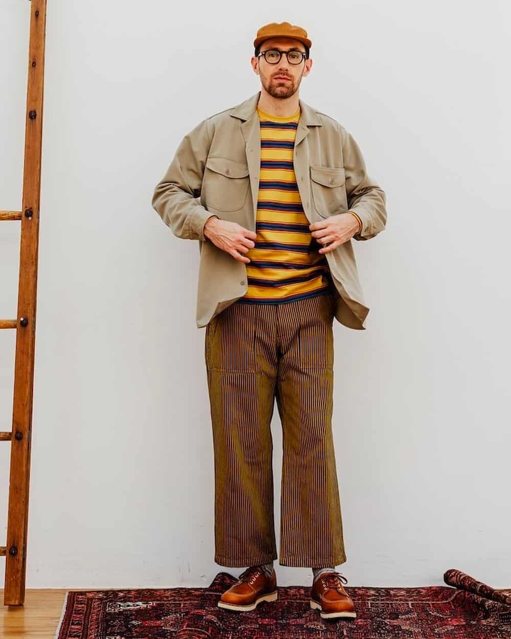 Man wearing Beam Plus wide leg brown pants, navy/yellow stripe T-shirt, khaki safari overshirt, tan leather shoes and tan baseball cap