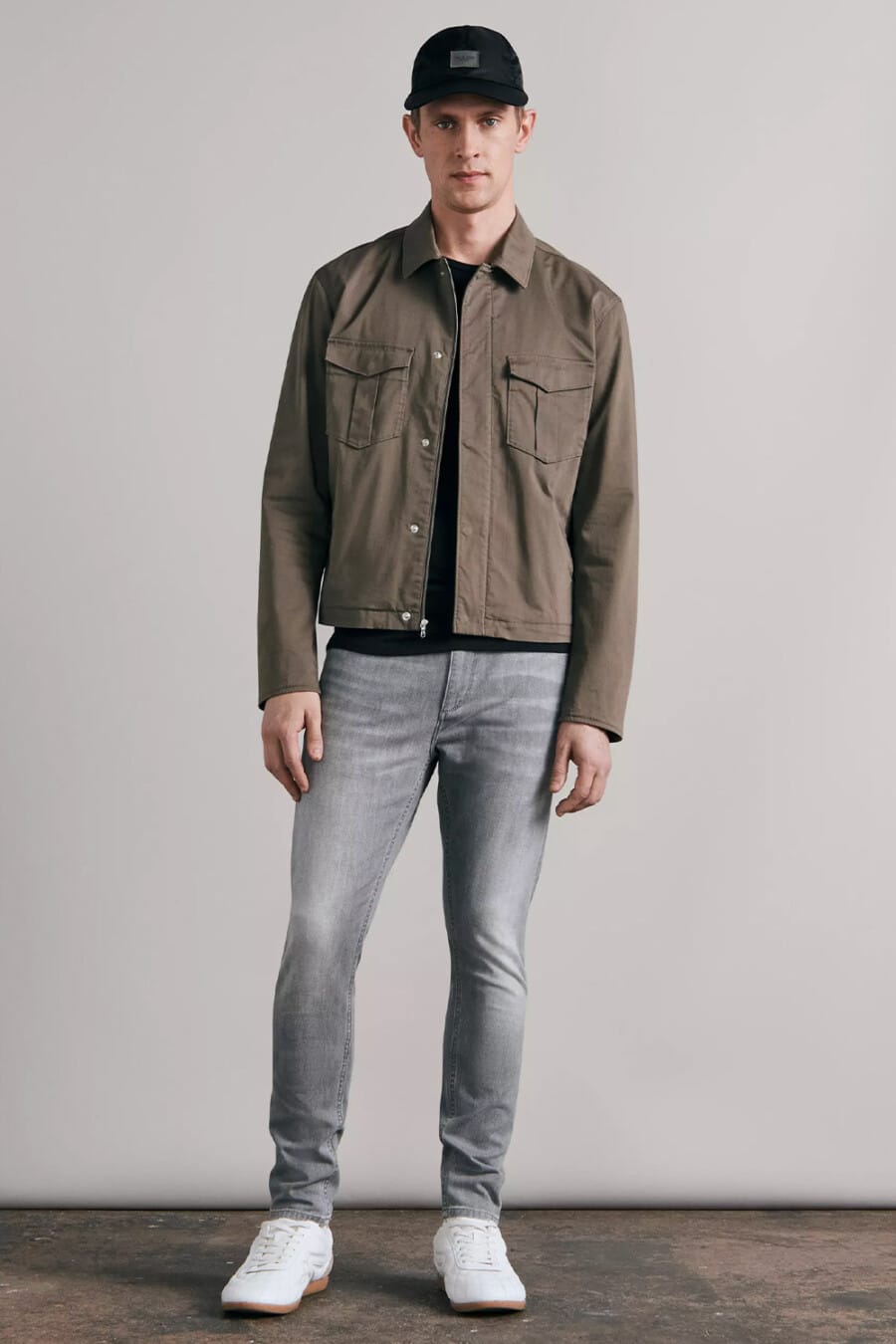 Men's light grey jeans, black T-shirt, olive green trucker jacket, black baseball cap and white sneakers outfit