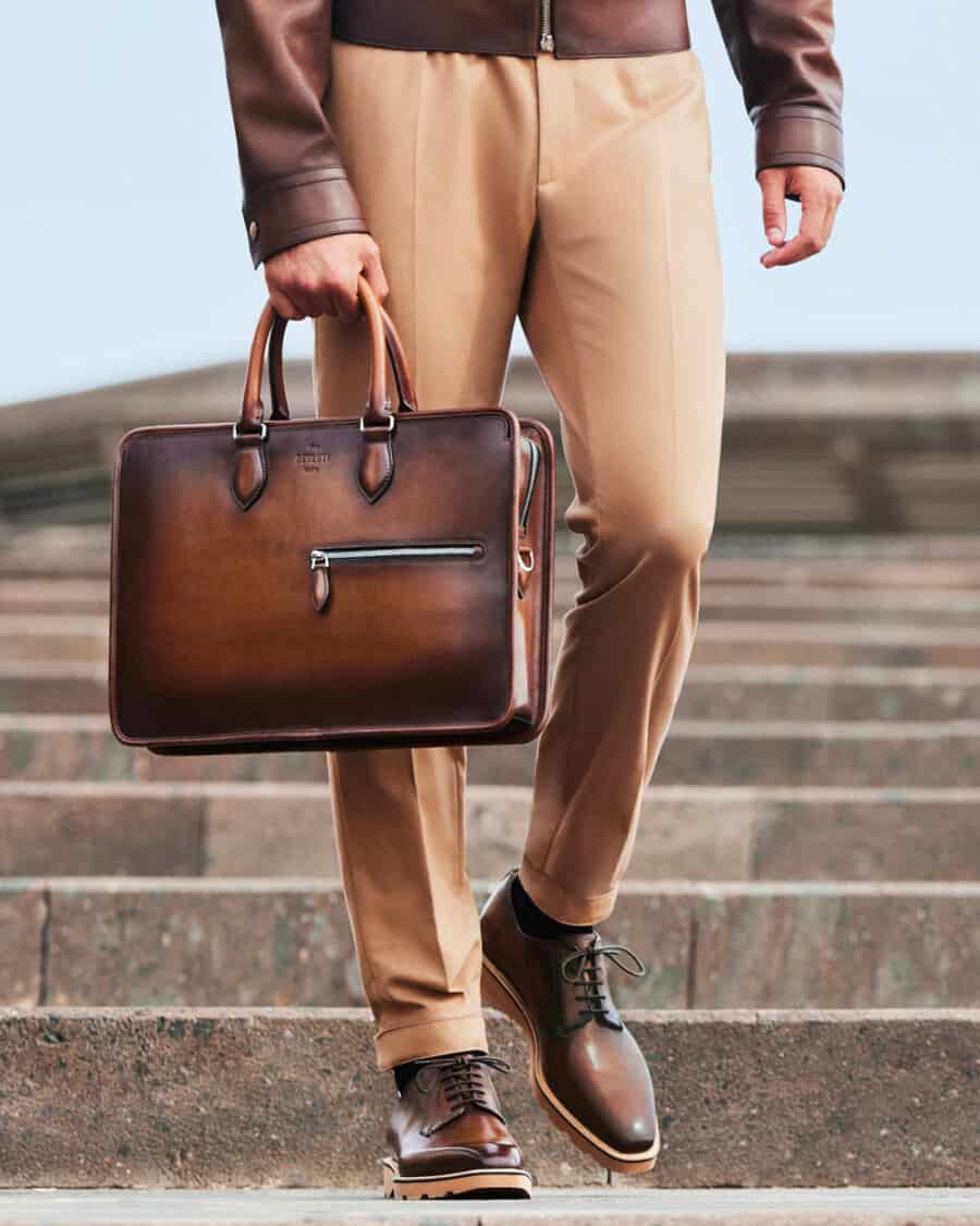 A man in camel tailored pants, brown leather jacket and brown leather Derby shoes carrying a luxury brown burnished leather briefcase by Berluti
