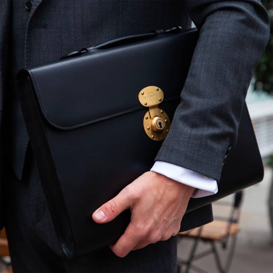 Man in a charcoal suit carrying a luxury black leather Ettinger briefcase with gold locking mechanism