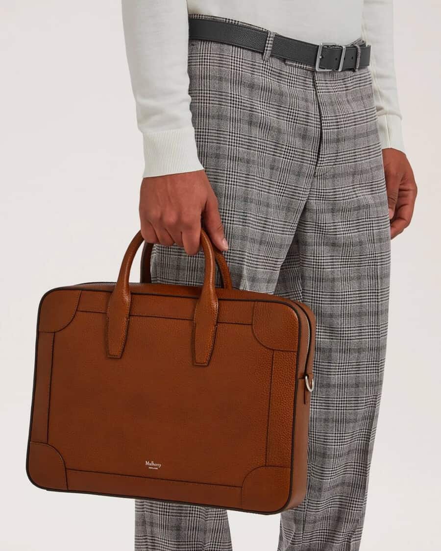 Man in grey plaid check pants holding a tan leather luxury Mulberry briefcase