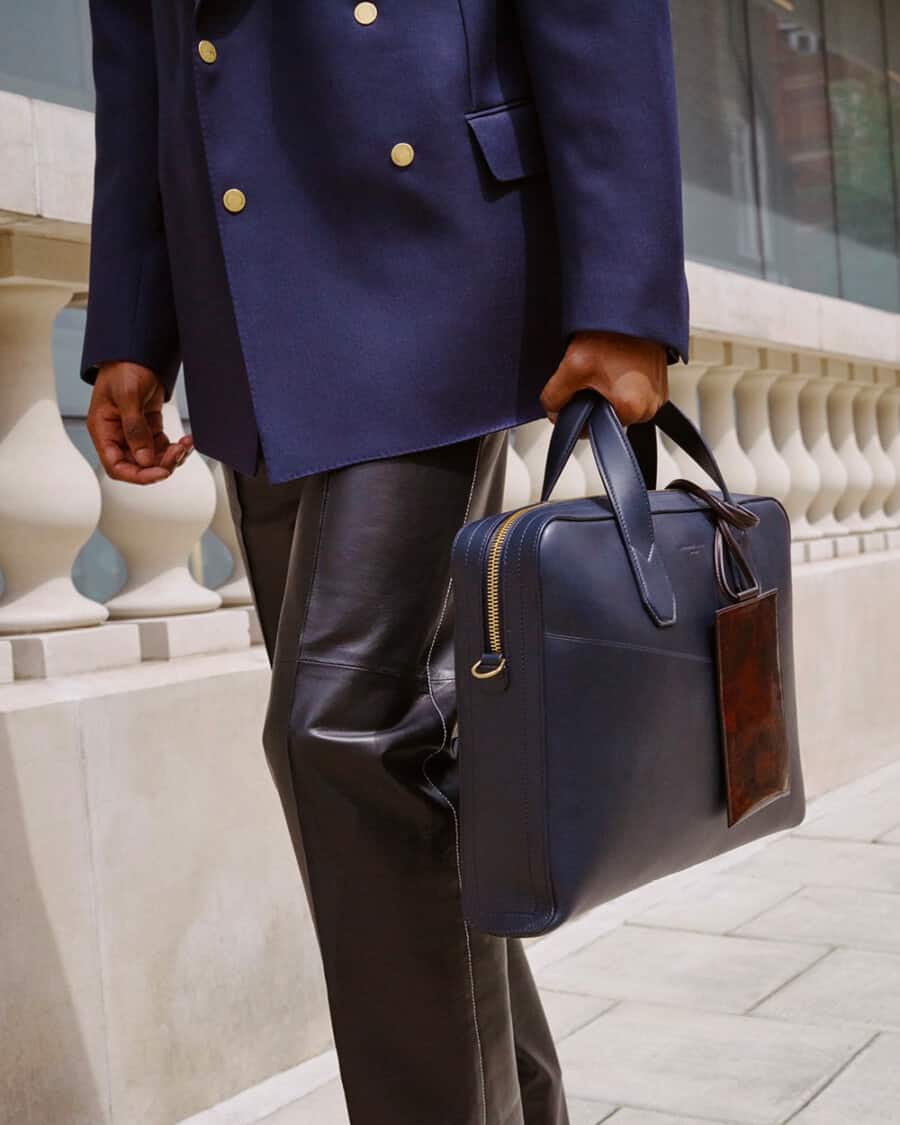 Man in black leather trousers and a navy gold button peacoat carry a luxury blue leather briefcase by Alfred Dunhill