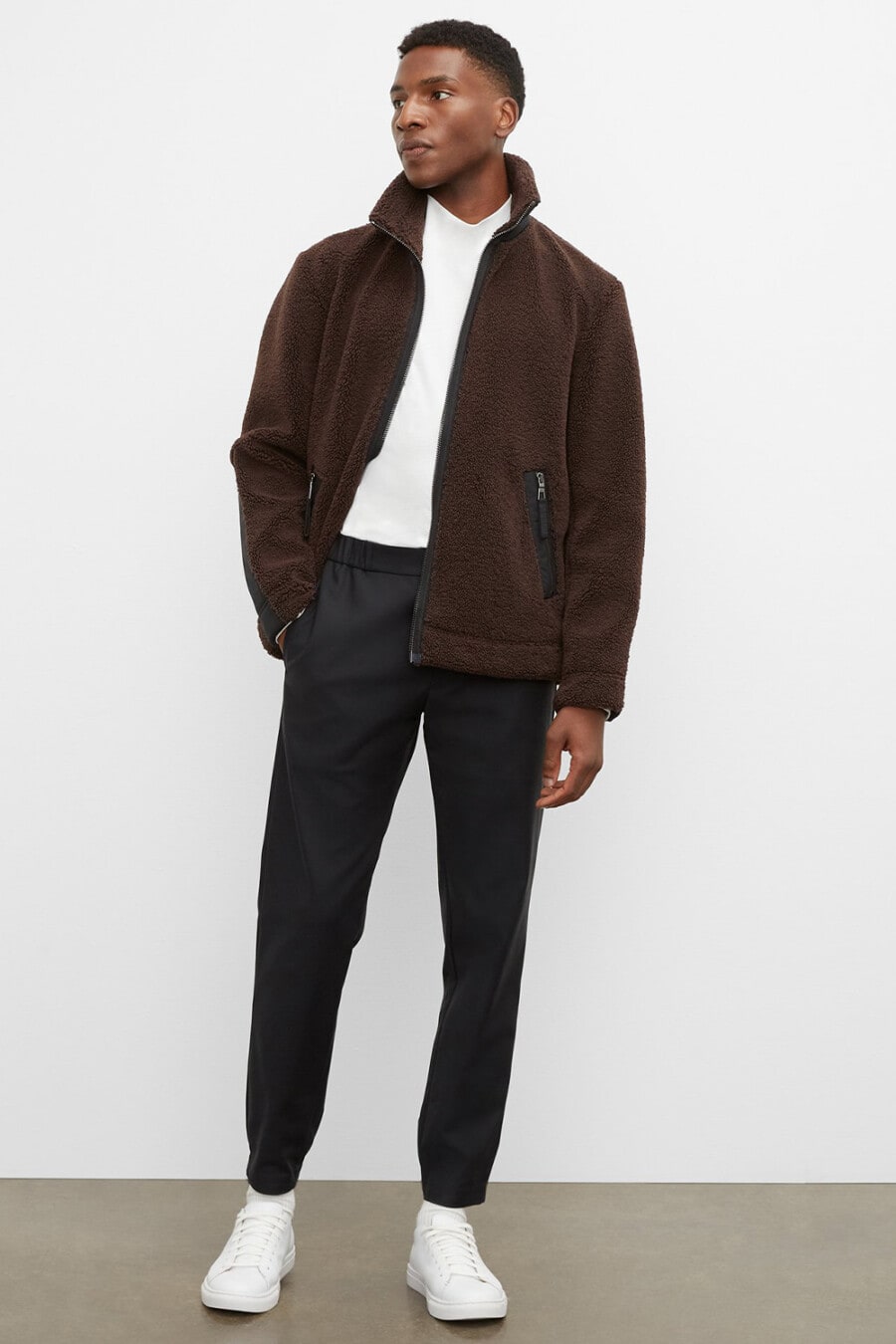 Men's black drawstring pants, tucked in white T-shirt, brown fleece jacket and white leather sneakers outfit