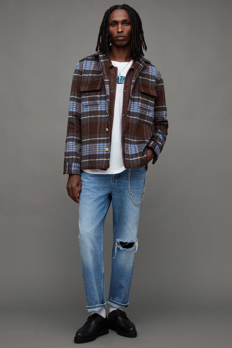 Men's ripped light wash jeans, white logo T-shirt, brown/blue checked overshirt, white socks, wallet chain and black monk-strap shoes grunge outfit