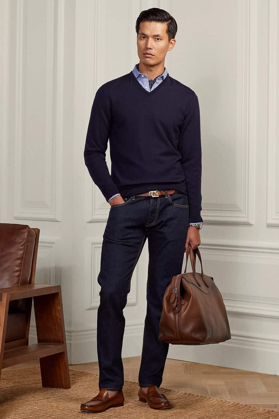 Men's dark raw jeans, light blue dress shirt, v-neck navy sweater, tan leather belt, tan penny loafers and brown weekender bag outfit