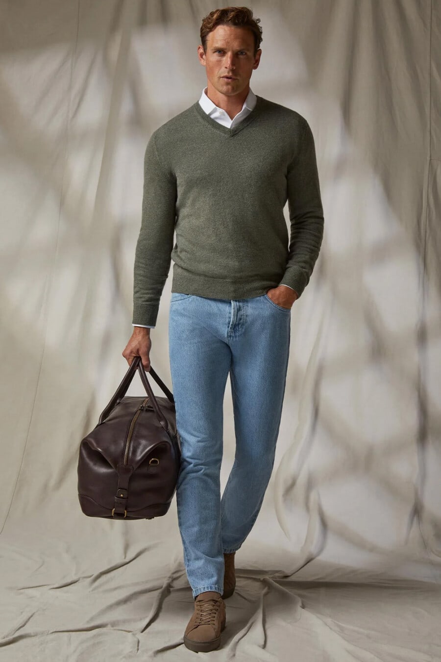 Men's pale light blue denim jeans, white dress shirt, green V-neck sweater, brown suede sneakers and brown leather weekender bag outfit