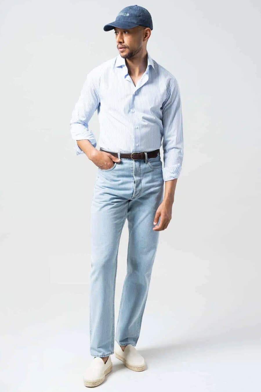 Men's light wash jeans, tucked in pale blue striped shirt, brown leather belt, white espadrilles and dark blue baseball cap outfit