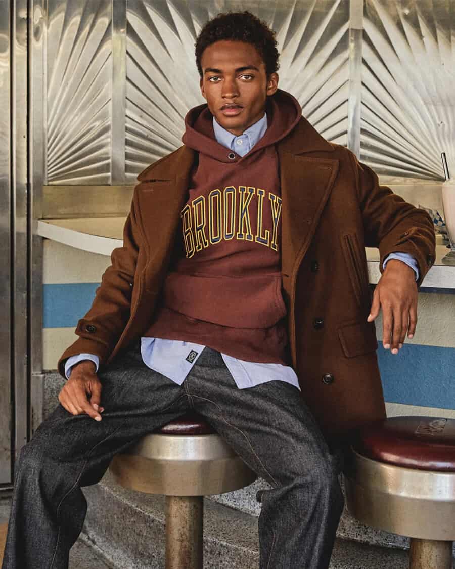 Man wearing Todd Snyder preppy burgundy Brooklyn hoodie with blue Oxford shirt, raw denim jeans and a brown overcoat