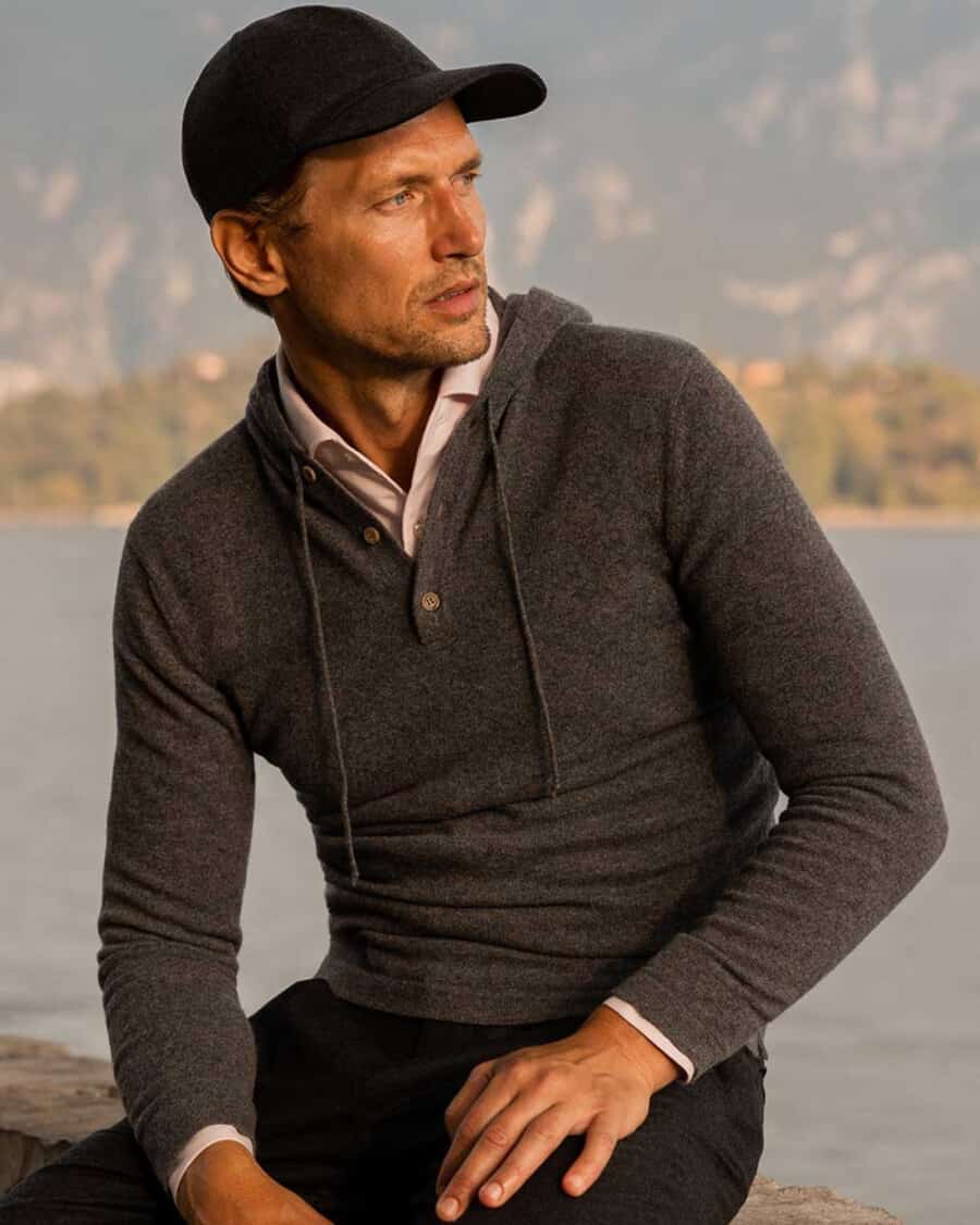 Man wearing a Luca Faloni charcoal cashmere quiet luxury hoodie over a pink shirt with black pants and a black premium baseball cap